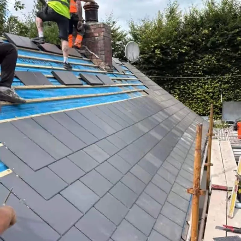 Roofers laying grey Thrutone slates over blue membrane and timber battens beside a chimney